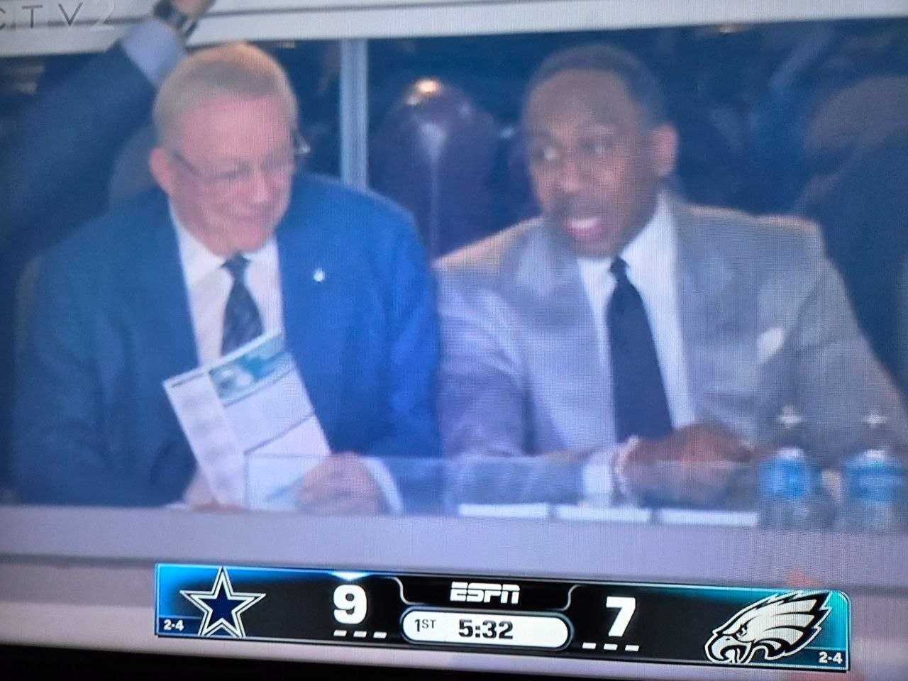 Photo of a TV showing ESPN's broadcast of the Dallas Cowboys vs Philadelphia Eagles game: Stephen A. Smith in a grey suit sits beside Cowboys owner Jerry Jones in a suite, with the score Cowboys 9, Eagles 7, 5:32 left in the first quarter.
